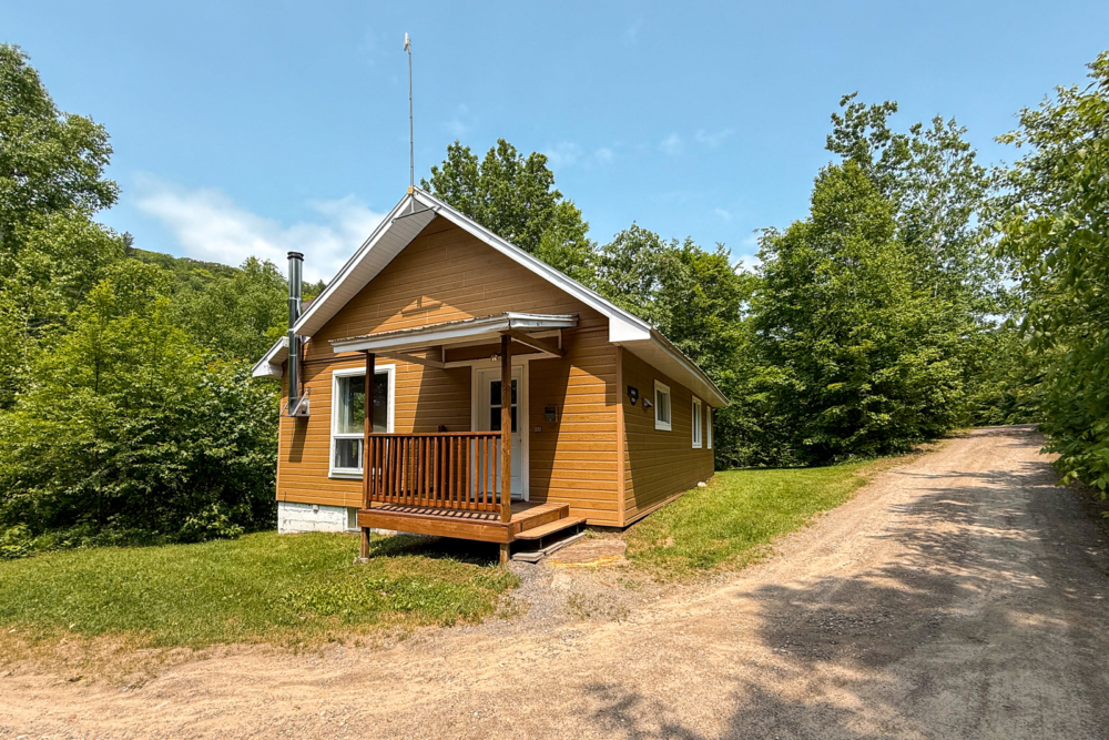 Chalet avec petit balcon couvert, entouré d’arbres et accessible par un chemin de gravier.