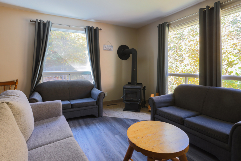 Salon chaleureux d’un chalet avec sofas, poêle à bois, grandes fenêtres et table basse ronde.