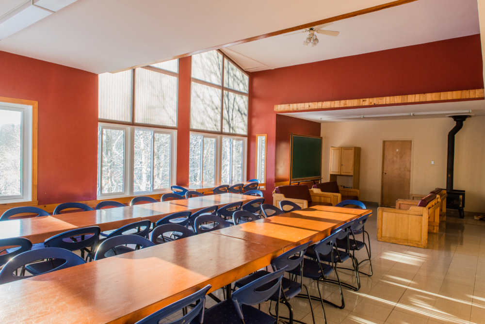 Salle à manger lumineuse d’un chalet avec grandes tables, chaises, fenêtres panoramiques et poêle à bois.