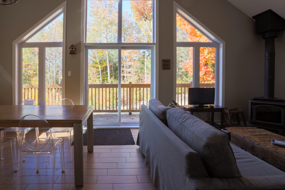 Intérieur chaleureux d’un chalet avec salon, salle à manger et grandes fenêtres donnant sur une forêt colorée d’automne.