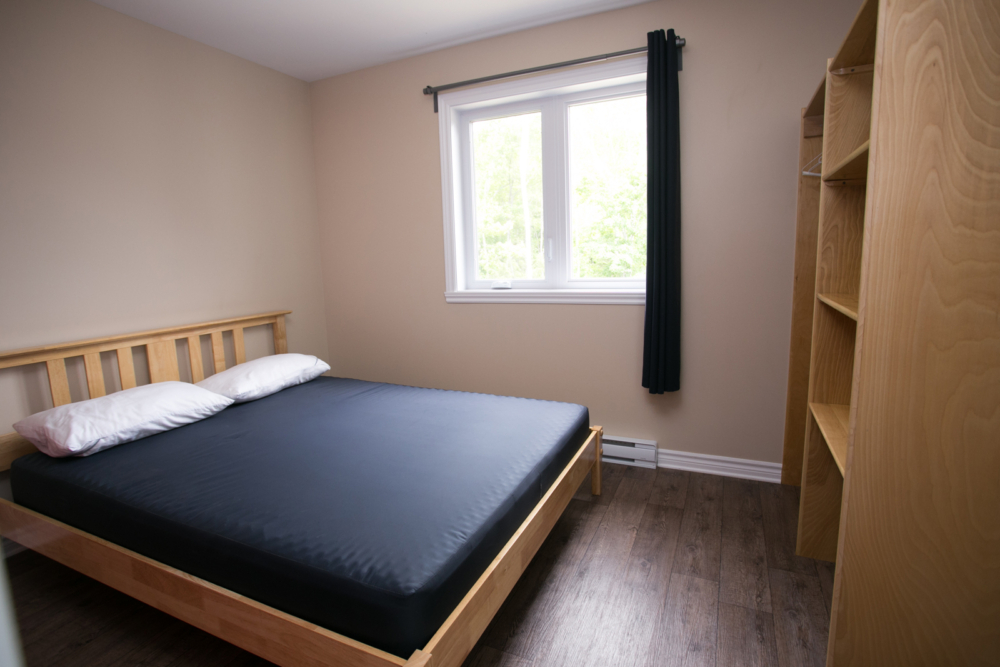 Chambre d’un chalet avec lit double, fenêtre lumineuse, rideaux noirs et rangement en bois.