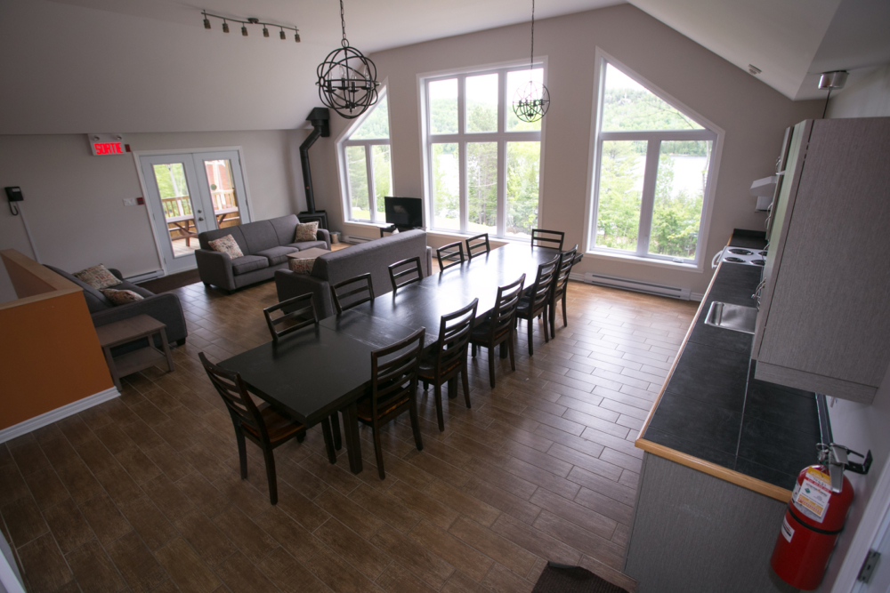 Intérieur lumineux d’un chalet avec grande salle à manger, cuisine, salon et vastes fenêtres donnant sur la forêt.