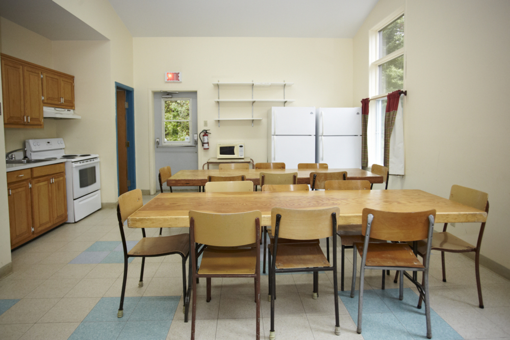 Cuisine et salle à manger communes d’un chalet, avec tables, chaises, électroménagers et grandes fenêtres.