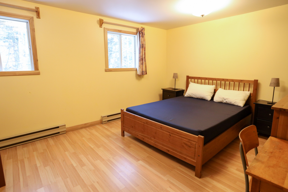 Chambre chaleureuse d’un chalet, avec lit double en bois, fenêtres lumineuses et mobilier en bois.
