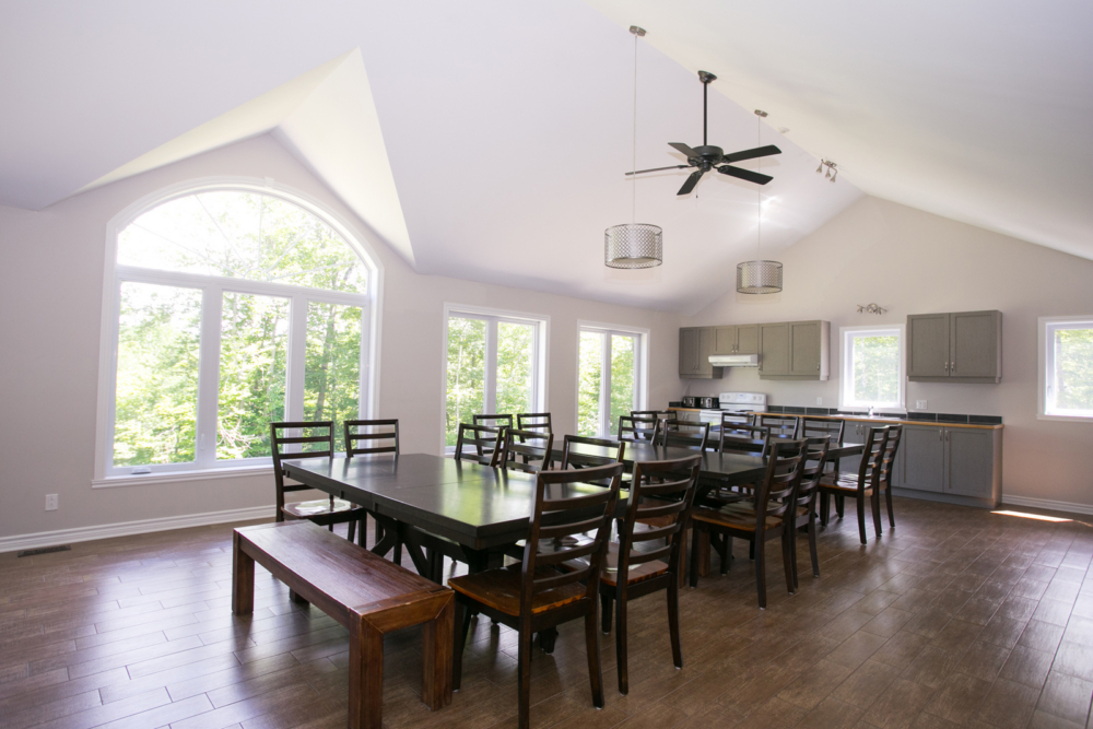 Grande salle à manger lumineuse d’un chalet, avec nombreuses places assises, cuisine équipée et fenêtres donnant sur la nature.