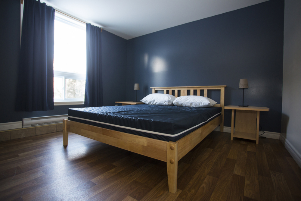 Chambre d’un chalet avec grand lit en bois, murs bleu foncé, tables de chevet et fenêtre donnant sur la nature.