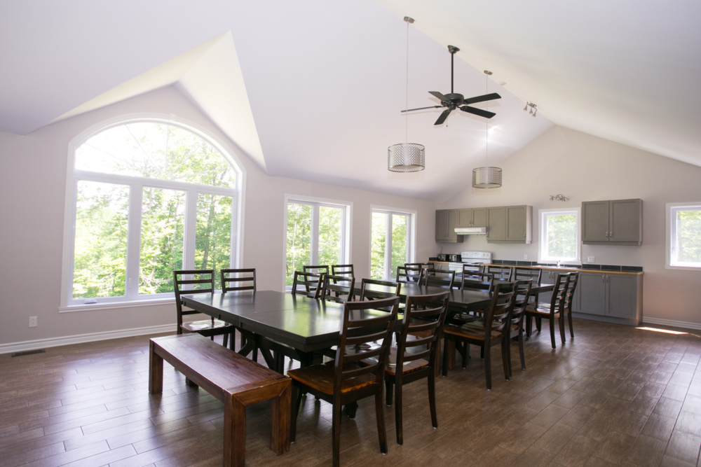 Grande salle à manger lumineuse d’un chalet, avec longues tables, nombreuses chaises et cuisine ouverte entièrement équipée.