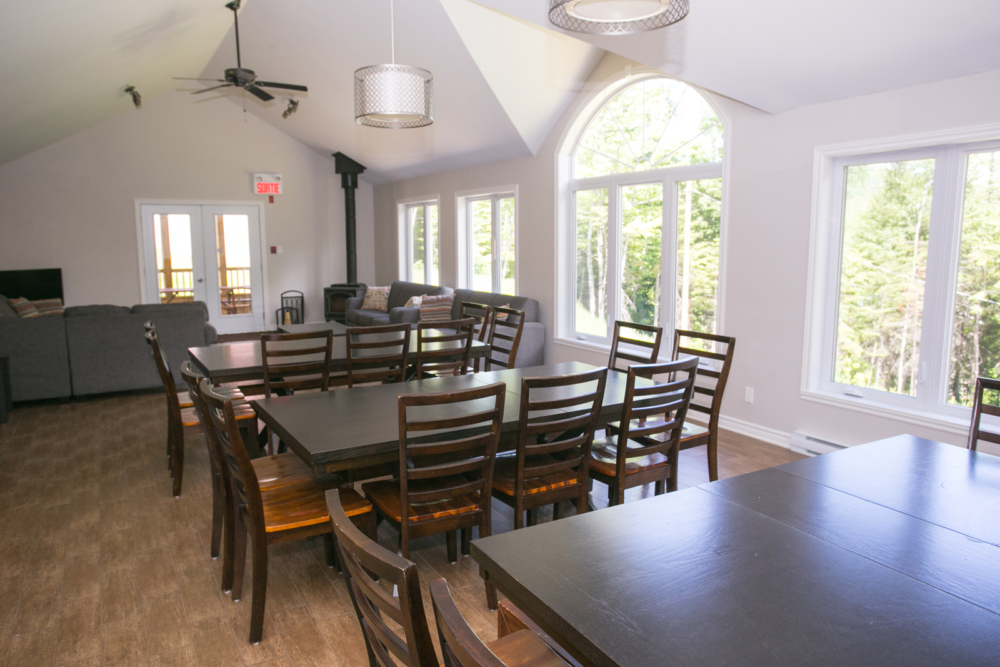 Salle à manger lumineuse d’un chalet avec grandes fenêtres, tables et nombreuses chaises en bois.
