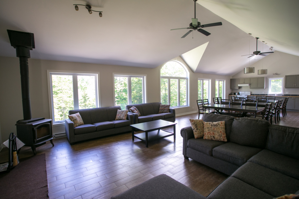 Intérieur spacieux d’un chalet avec salon, poêle à bois, grandes fenêtres et cuisine ouverte.