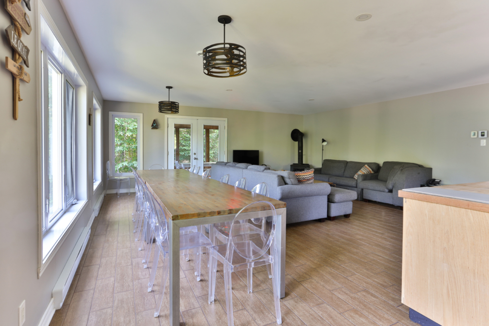 Intérieur lumineux d’un chalet avec grande table à manger, chaises transparentes, sofas gris et poêle à bois.