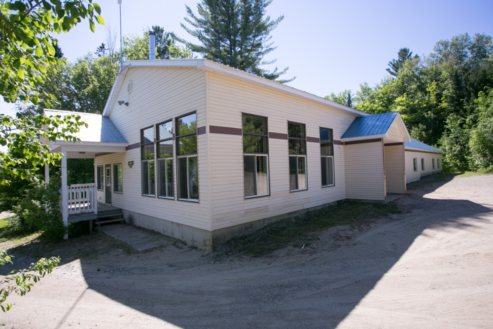 Chalet avec grandes fenêtres, terrasse couverte et entrée en gravier, entouré d’arbres verdoyants.