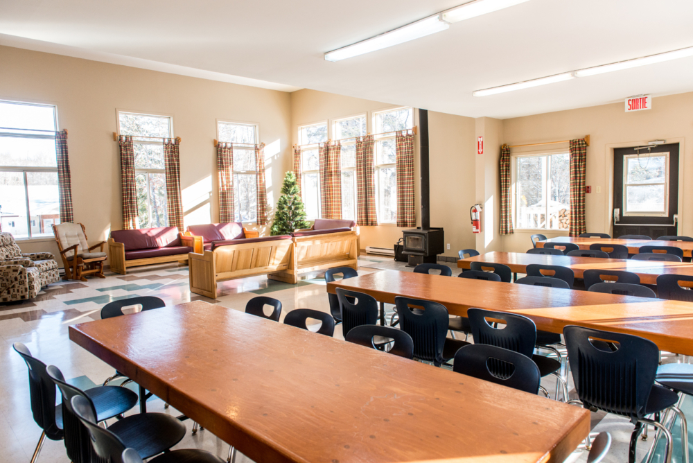 Salle commune lumineuse d’un chalet avec grandes tables, chaises, canapés et foyer au bois.