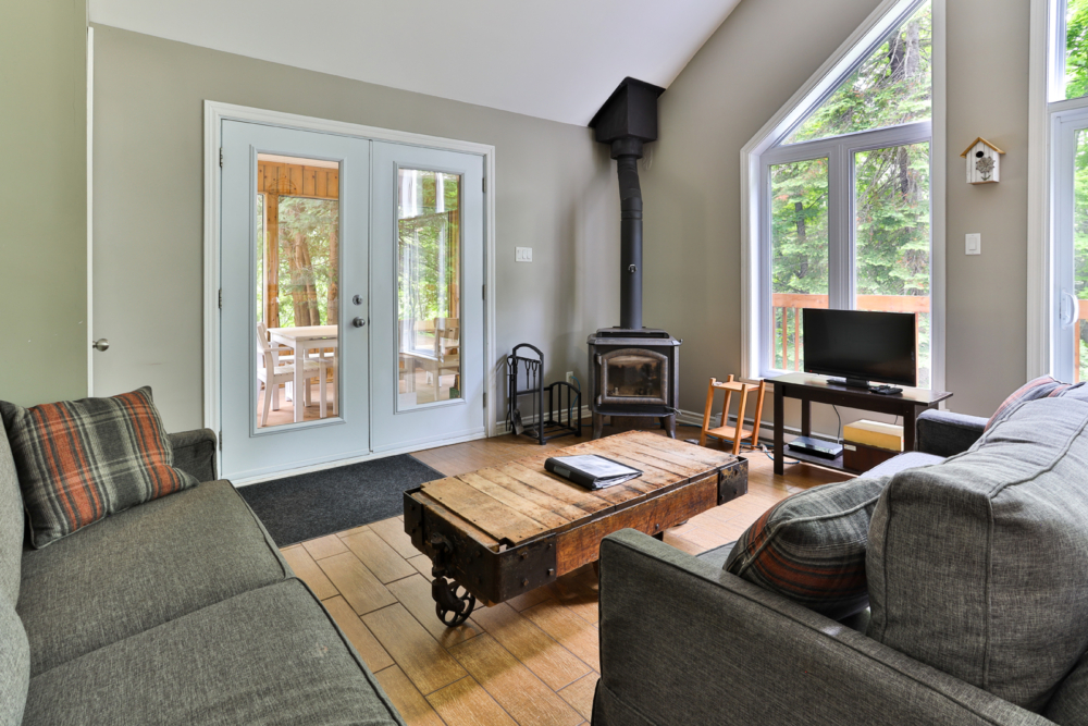 Salon chaleureux d’un chalet avec poêle à bois, sofas, table basse rustique et grandes fenêtres donnant sur la forêt.