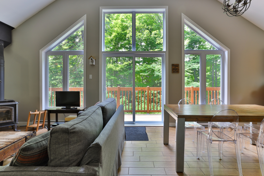 Intérieur lumineux d’un chalet avec salon, poêle à bois, salle à manger et grandes fenêtres donnant sur la forêt.