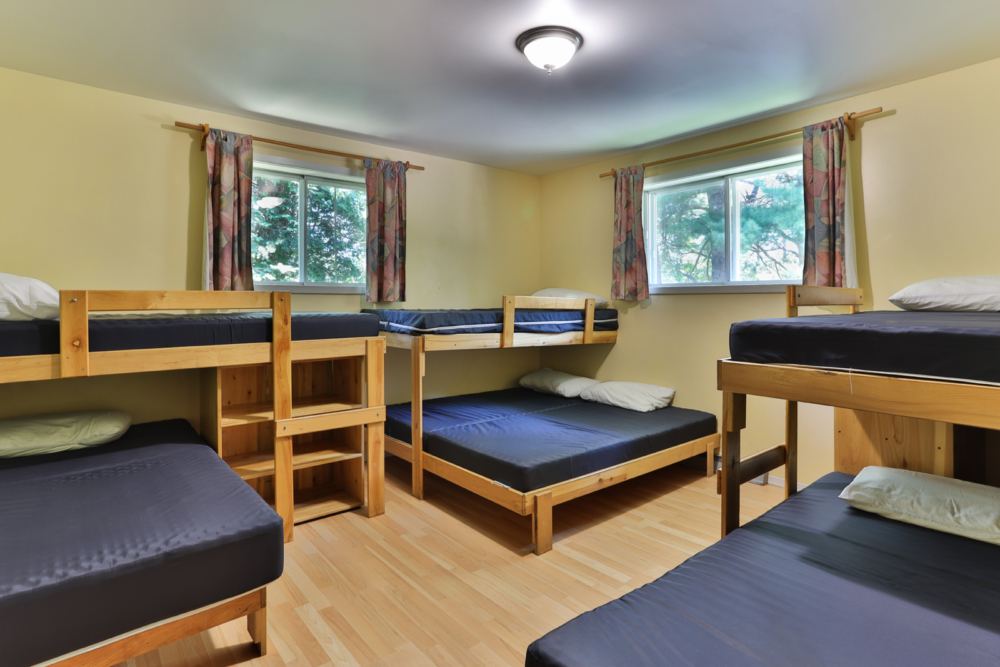 Chambre de chalet avec plusieurs lits superposés et lits simples, fenêtres donnant sur les arbres et mobilier en bois.