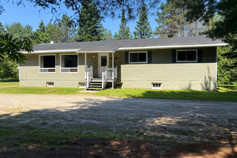 Cottage surrounded by trees, with a covered entrance, large windows and a spacious gravel driveway.