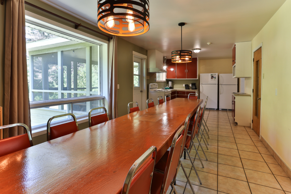Grande salle à manger d’un chalet, avec longue table en bois, nombreuses chaises et cuisine équipée.