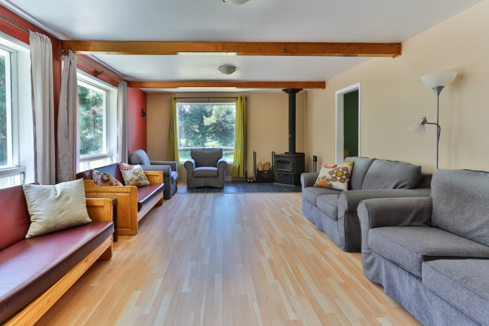 Salon chaleureux d’un chalet, avec canapés, fauteuils, poêle à bois et grandes fenêtres donnant sur la forêt.
