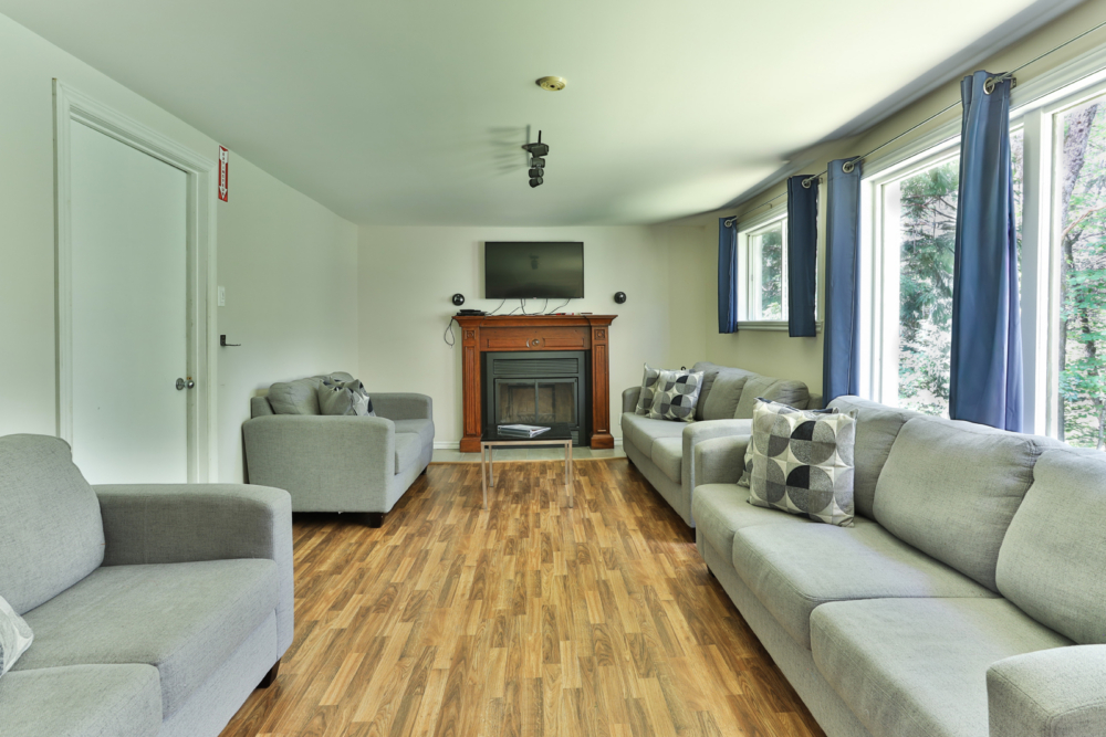 Salon chaleureux d’un chalet avec foy­er, téléviseur, canapés confortables et grandes fenêtres donnant sur la nature.