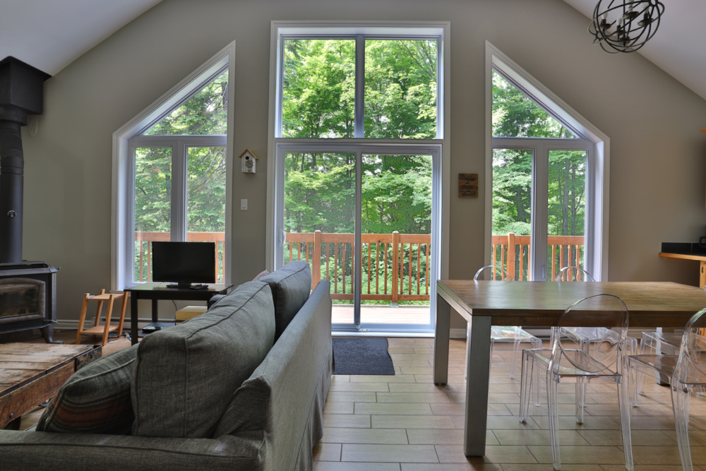 Intérieur lumineux d’un chalet avec grandes fenêtres, vue sur la forêt, salon, poêle à bois et salle à manger.