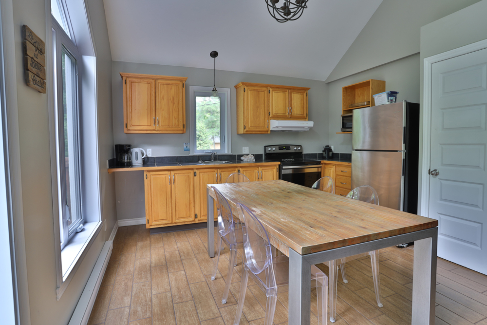 Cuisine d’un chalet avec armoires en bois, grandes fenêtres, table à manger et électroménagers.