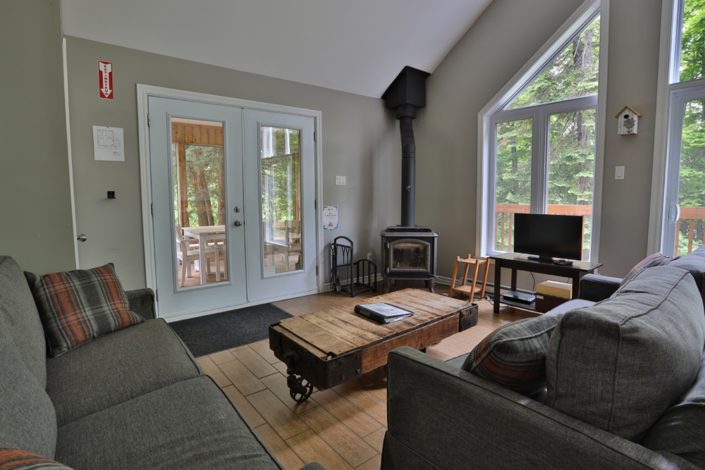 Salon chaleureux d’un chalet avec divans, poêle à bois, téléviseur et grandes fenêtres donnant sur la forêt.
