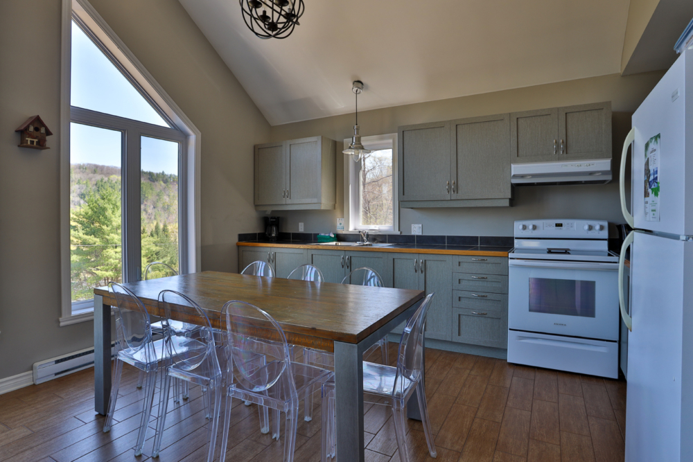Cuisine lumineuse d’un chalet, avec table à manger, chaises transparentes et vue sur la forêt.