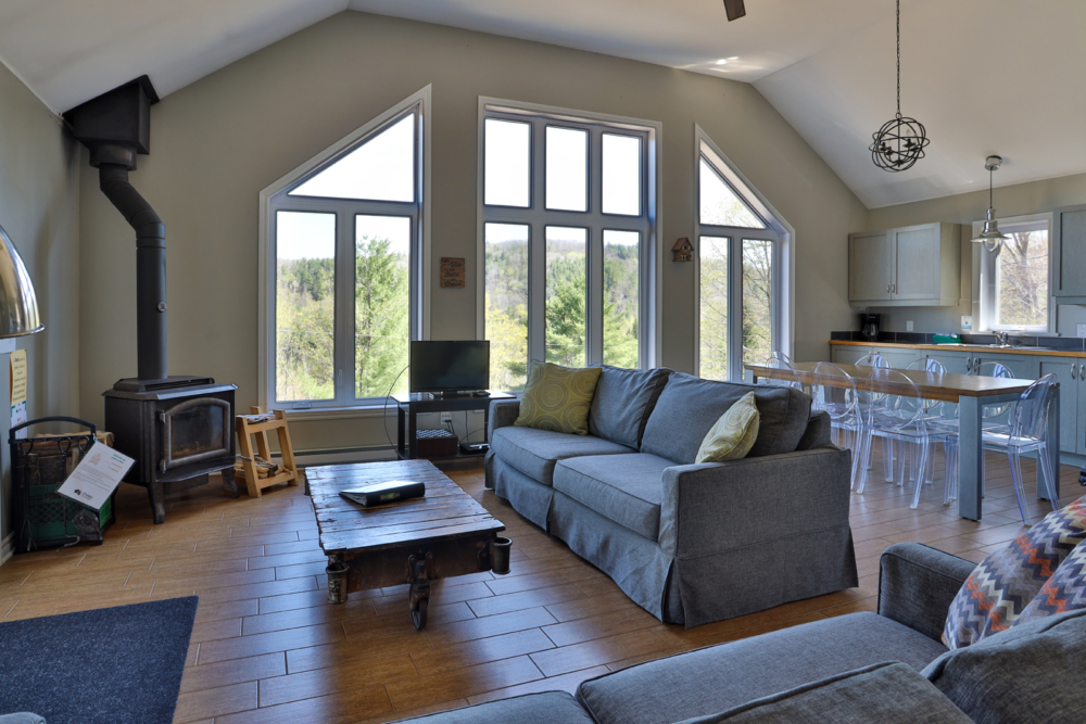 Intérieur chaleureux d’un chalet avec poêle à bois, grandes fenêtres, salon et cuisine ouverte.