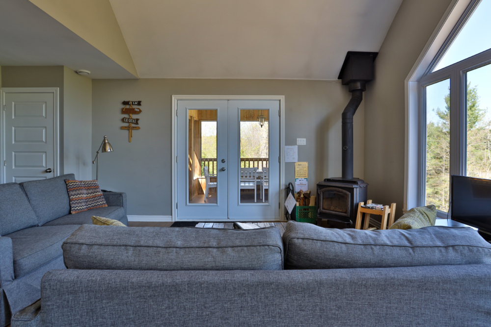 Salon chaleureux d’un chalet avec poêle à bois, grandes fenêtres et accès à la terrasse.