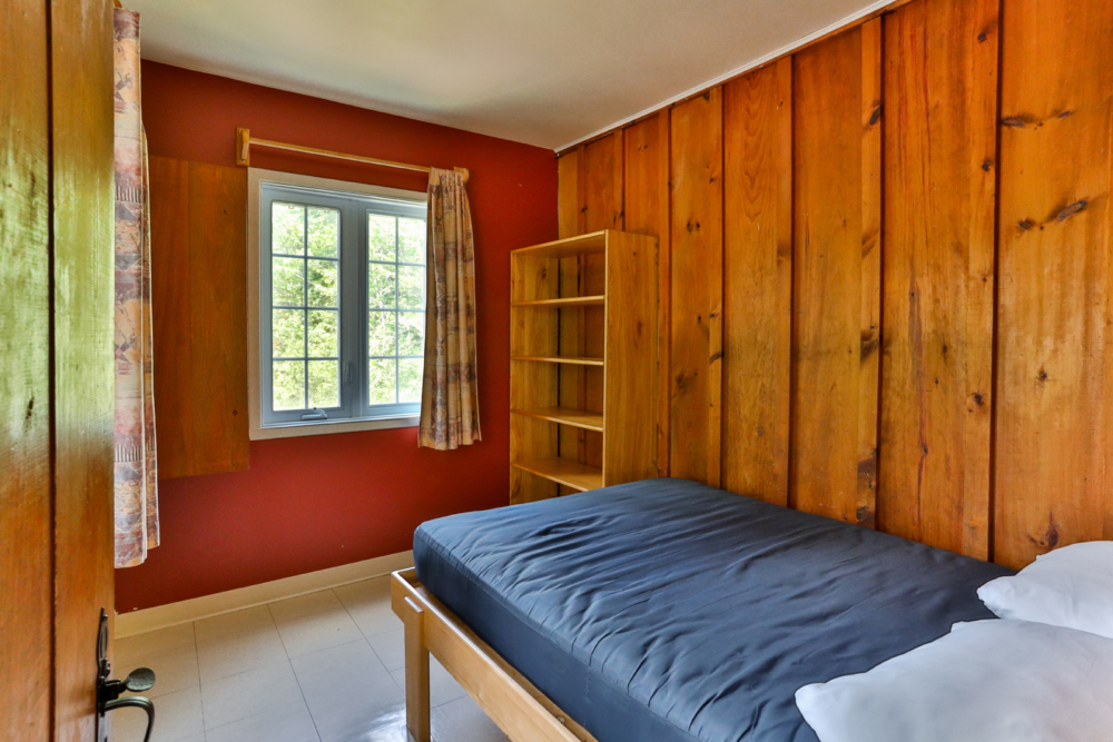 Chambre chaleureuse d’un chalet avec lit double, murs en bois, étagère et fenêtre donnant sur la nature.
