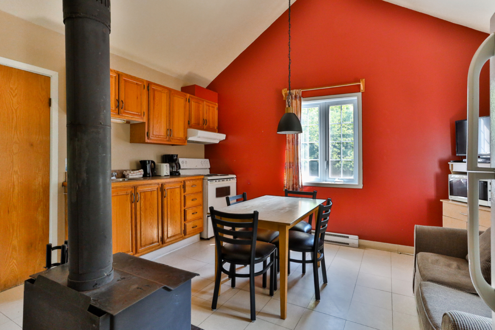 Cuisine-salle à manger chaleureuse d’un chalet aux murs rouges, avec poêle à bois, table et quatre chaises.