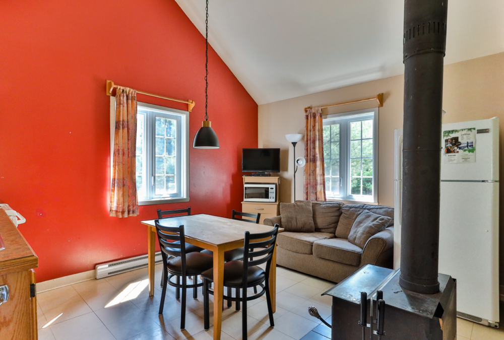 Intérieur chaleureux d’un chalet avec cuisine, salle à manger, salon, poêle à bois et grandes fenêtres.