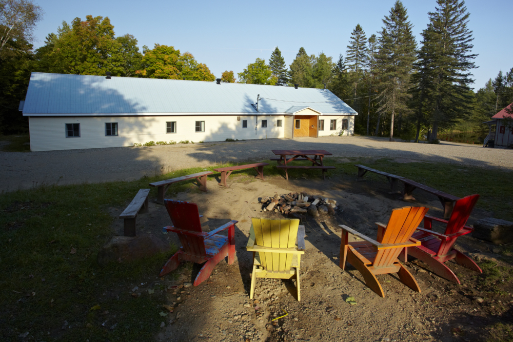 Aire de feu de camp avec chaises Adirondack devant un chalet, entouré d’arbres.