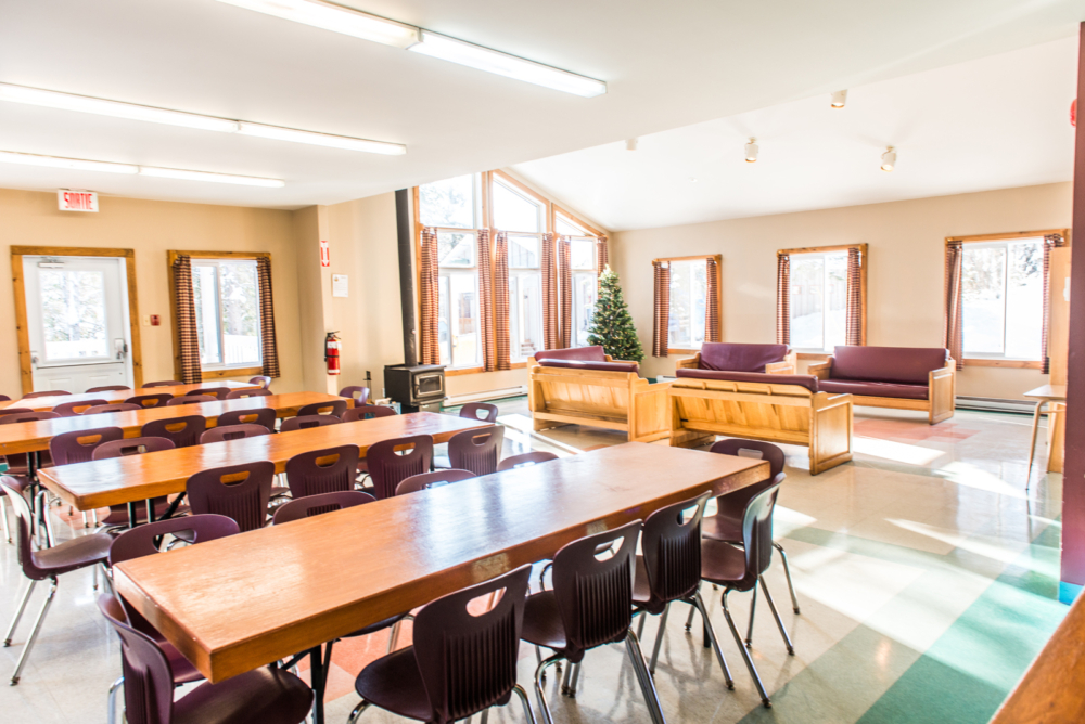Grande salle commune lumineuse d'un chalet avec tables et chaises, canapés, fenêtres panoramiques et sapin de Noel.