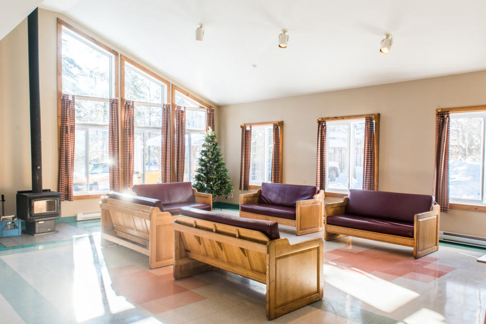 Salon lumineux d’un chalet avec grandes fenêtres, fauteuils en bois, poêle à bois et sapin de Noël.