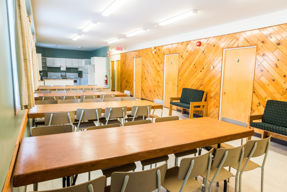 Salle à manger d'un chalet avec tables, chaises, fauteuils et espace cuisine, idéale pour les groupes.