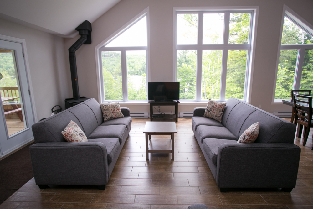 Salon lumineux d’un chalet avec deux canapés, foyer, téléviseur et grandes fenêtres donnant sur la nature.