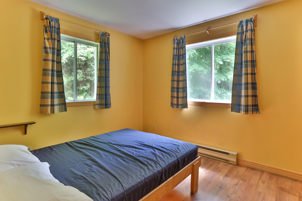 Chambre chaleureuse d’un chalet avec lit double, murs jaunes, rideaux à carreaux et vue sur la forêt.