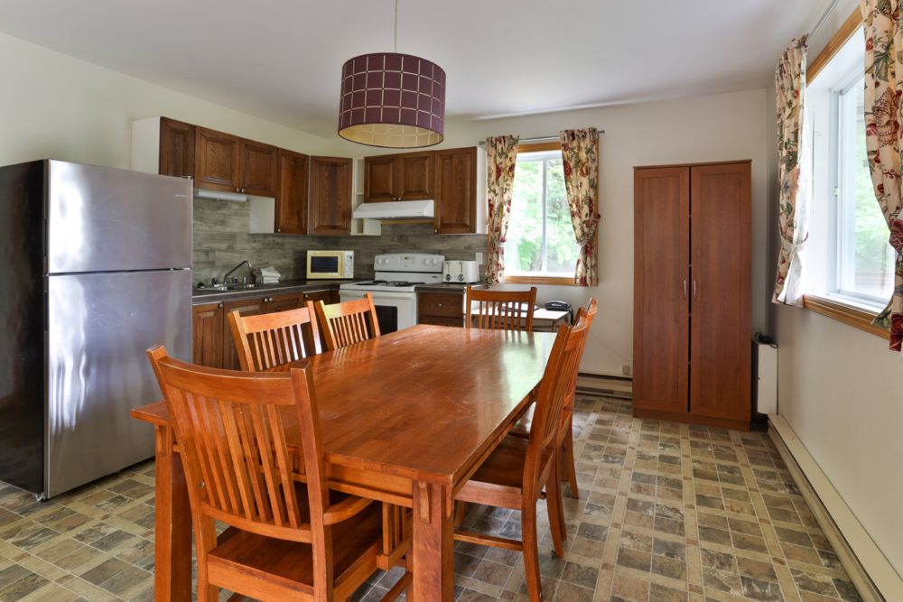 Cuisine entièrement équipée d’un chalet, avec grande table à manger en bois, chaises, électroménagers et fenêtres donnant sur la nature.