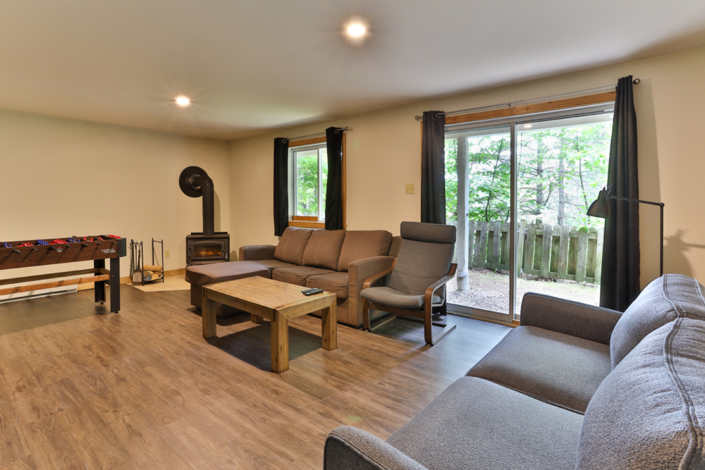 Salon chaleureux d’un chalet avec canapé, poêle à bois, table basse et grandes portes vitrées donnant sur la forêt.