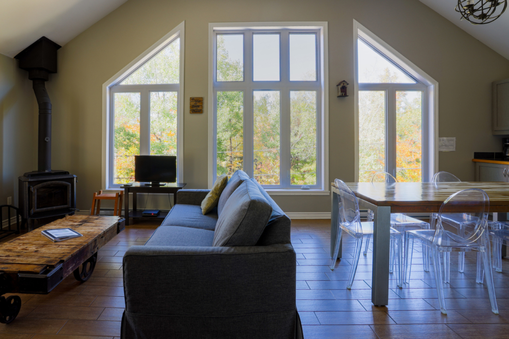 Intérieur lumineux d’un chalet avec salon, poêle à bois, grandes fenêtres, table à manger et vue sur la forêt.