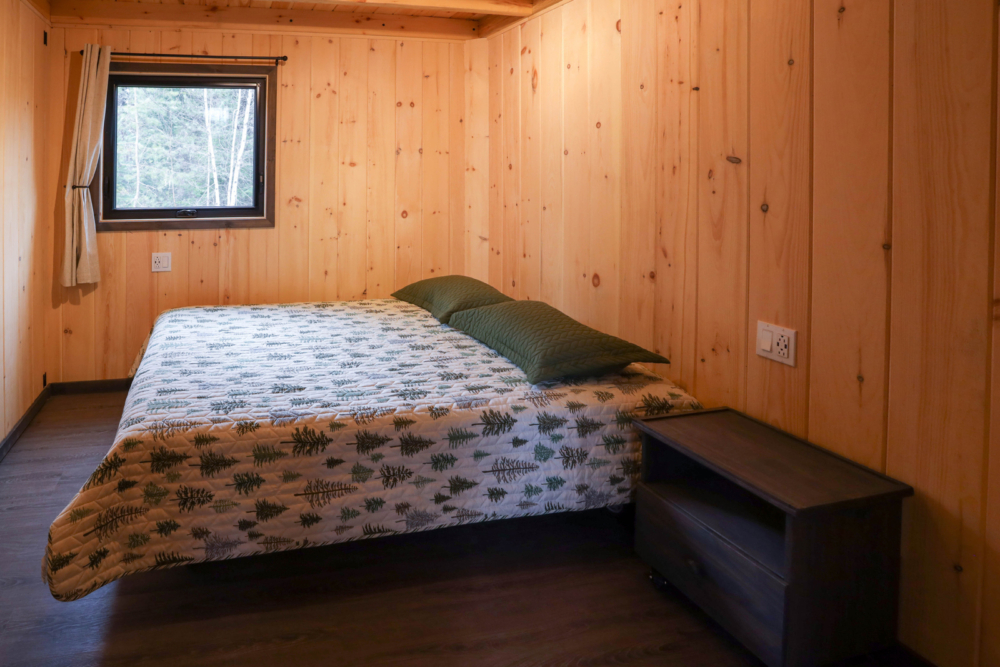 Chambre chaleureuse d’un mini-chalet en bois avec lit double, literie, tables de chevet et fenêtre donnant sur la forêt.
