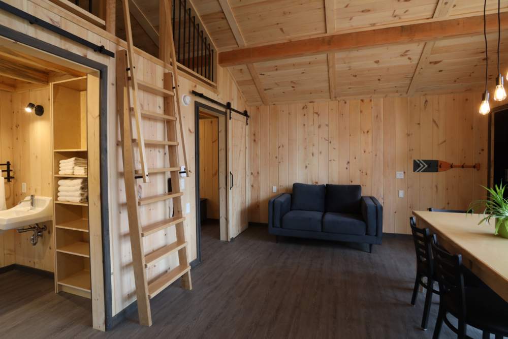 Intérieur chaleureux d’un mini-chalet avec canapé, espace repas, échelle en bois et salle de bain ouverte.