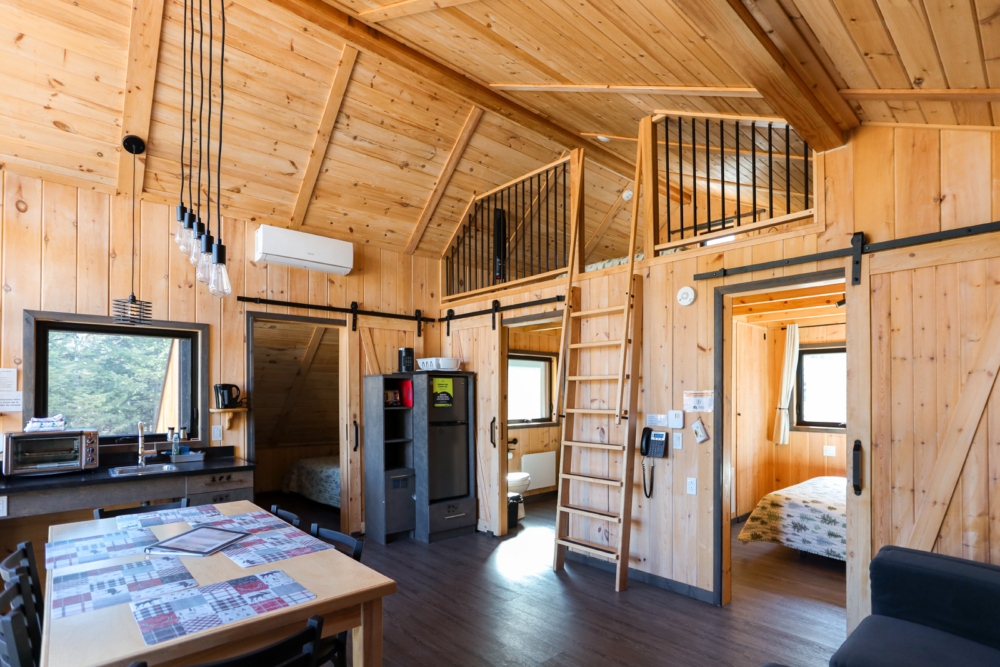 Intérieur chaleureux d’un mini-chalet en bois avec mezzanine, cuisine, table à manger et chambre.
