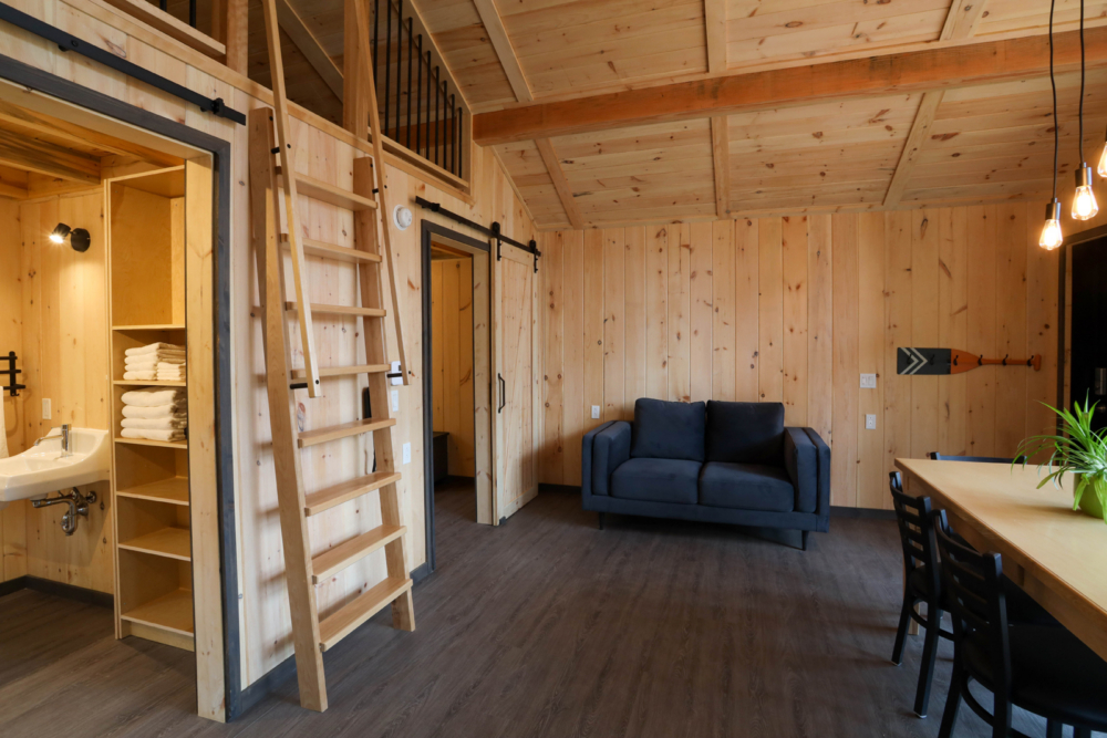 Intérieur chaleureux d’un mini-chalet avec canapé, table à manger, mezzanine et salle de bain visible.