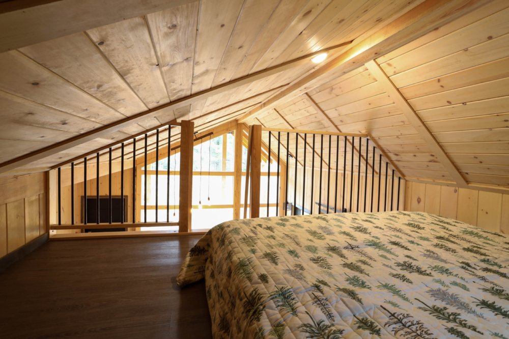 Mezzanine chaleureuse d’un mini-chalet avec lit double, plafond en bois et vue sur la pièce principale.