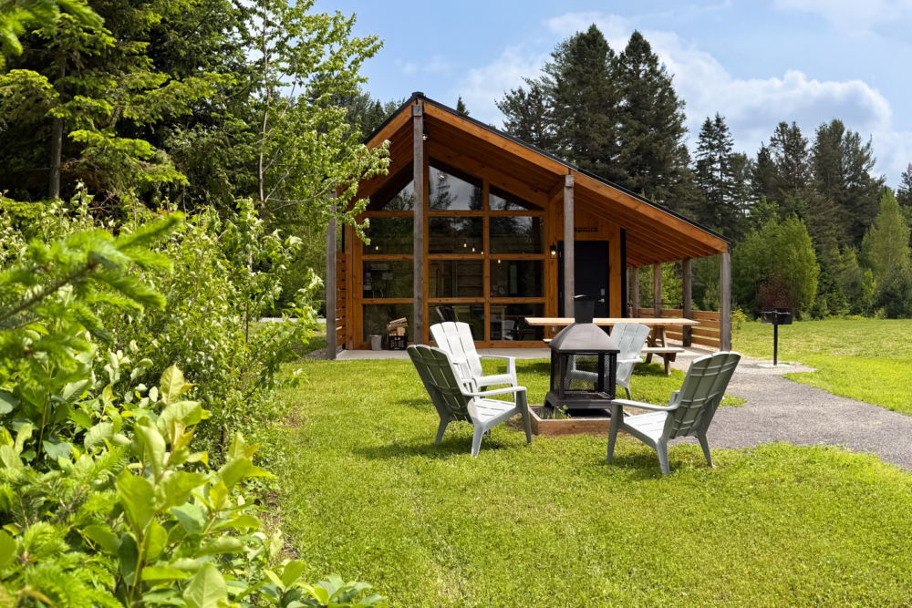Mini-chalet moderne en bois fenêtré, entouré d’arbres, foyer extérieur et chaises Adirondack sur la pelouse.