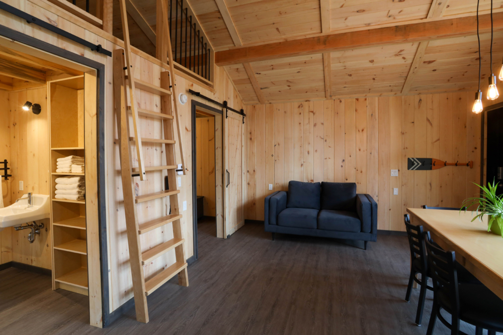 Intérieur chaleureux d’un mini-chalet avec salon, mezzanine, échelle en bois et espace repas.