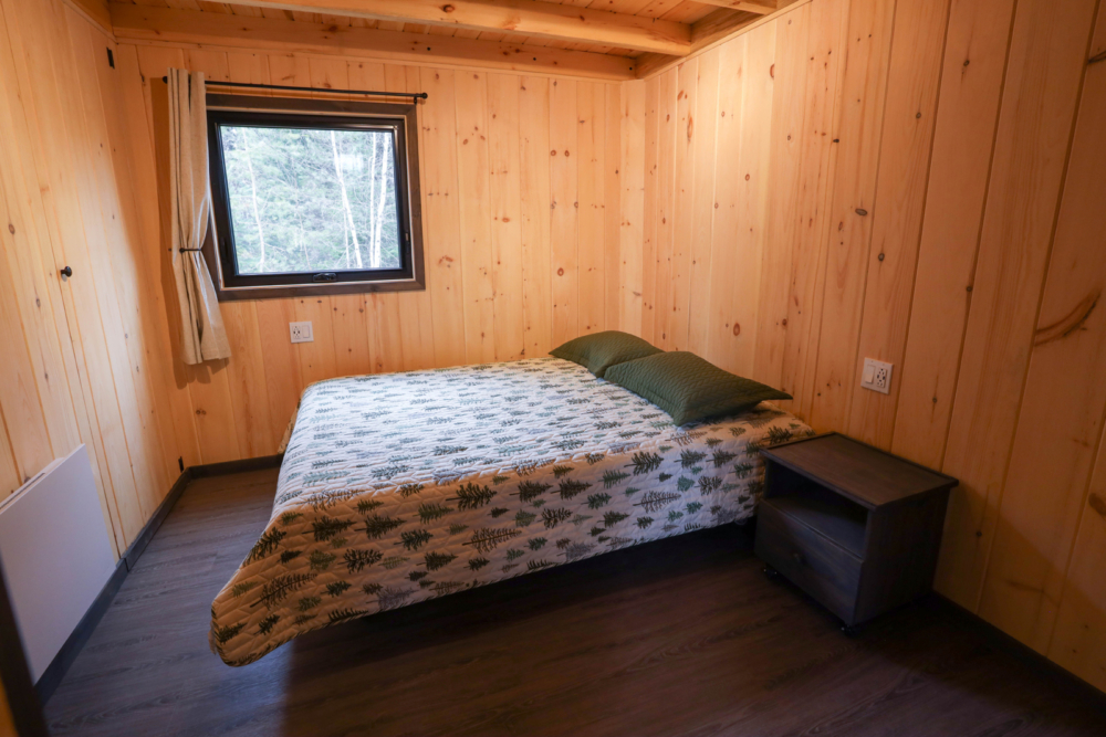 Chambre confortable d’un mini-chalet en bois avec lit double, literie à motif et fenêtre donnant sur la forêt.