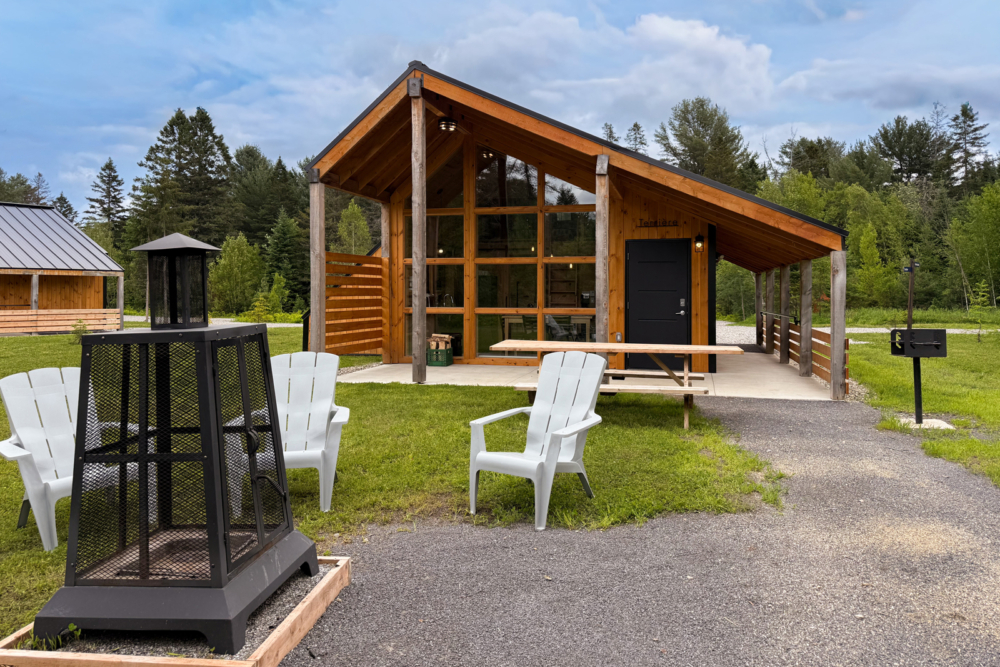 Mini-chalet en bois entouré d’arbres, sous un ciel bleu parsemé de nuages. Foyer extérieur et chaises.